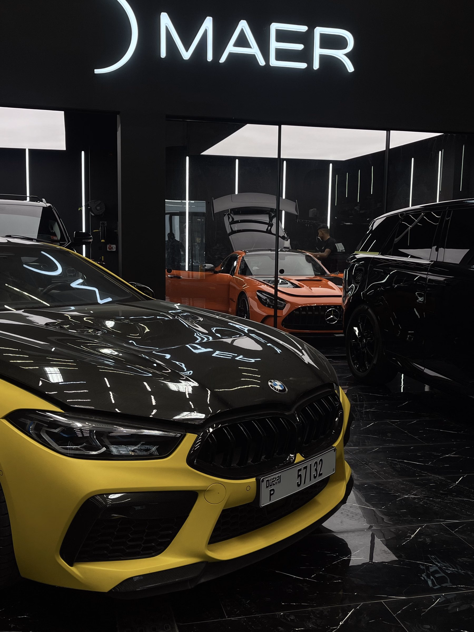 A yellow BMW M8 with a carbon fibre bonnet photographed from the front beneath the MAER sign after full body paint protection film