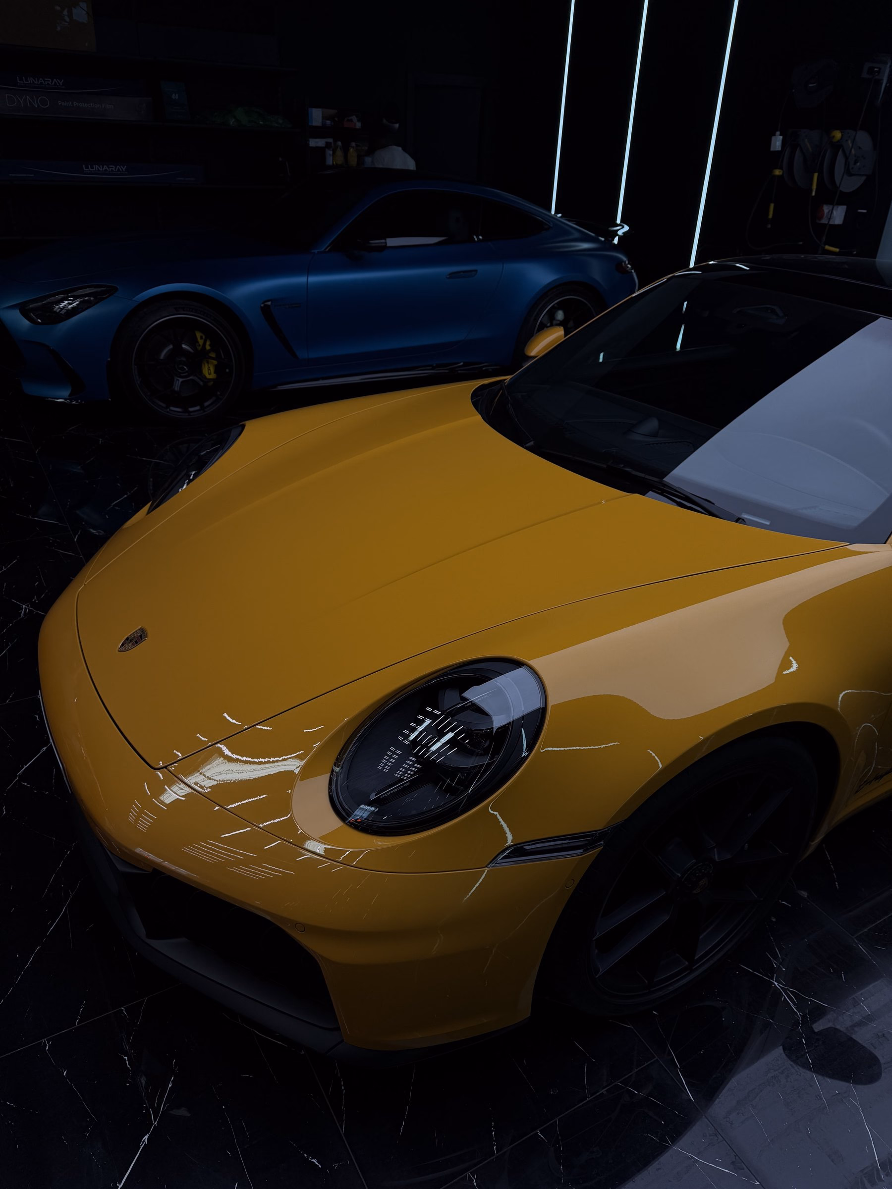 Front of a yellow Porsche 911 Turbo protected with full body paint protection film, a blue Mercedes-AMG behind it on the workshop floor
