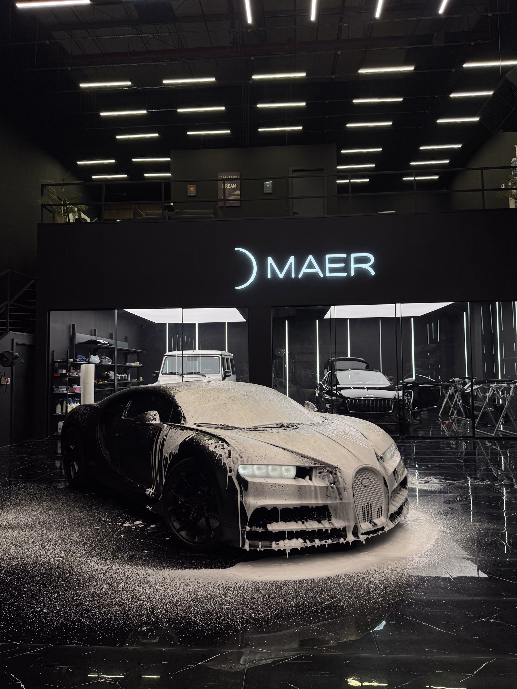 Bugatti covered in cleaning foam under studio lighting, the illuminated MAER sign visible behind it