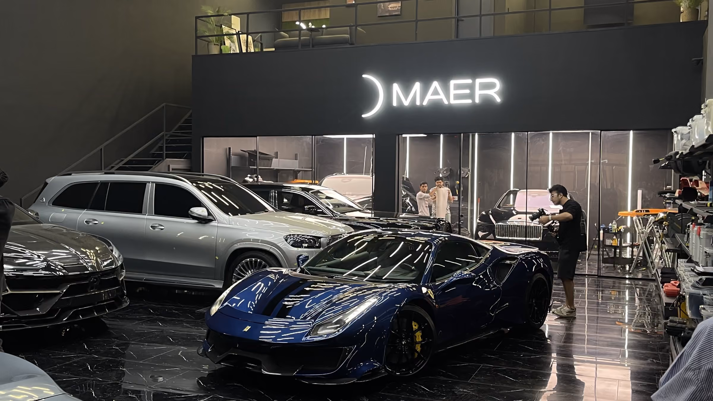 The MAER workshop floor in Al Quoz with a Ferrari 488, a Lamborghini Urus and a Mercedes GLS under the illuminated MAER sign while staff work